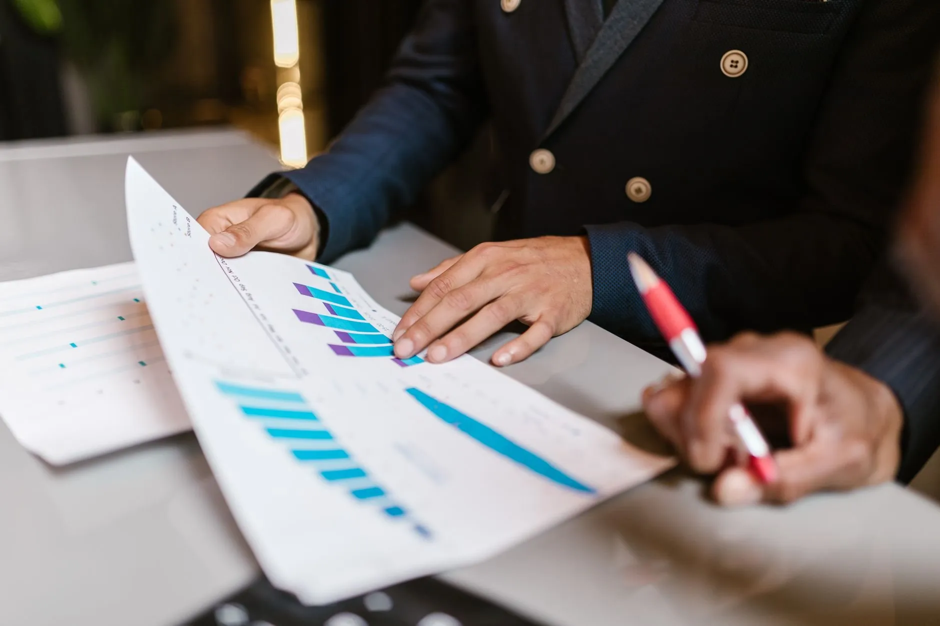 Two people in suits reviewing printed bar charts at a meeting table, one holding a red pen