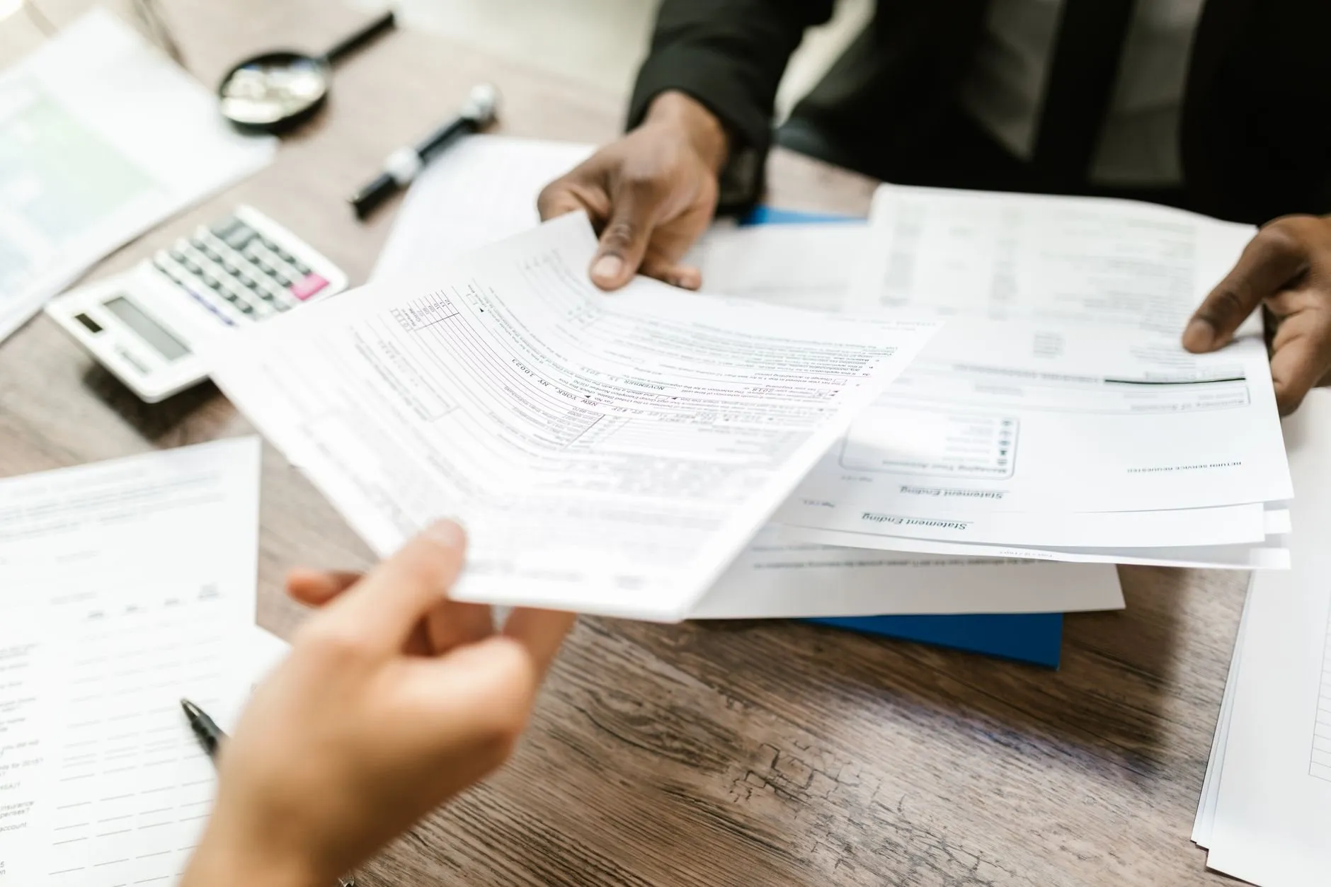 One person handing printed financial forms across a wooden desk to another, a calculator and magnifier nearby