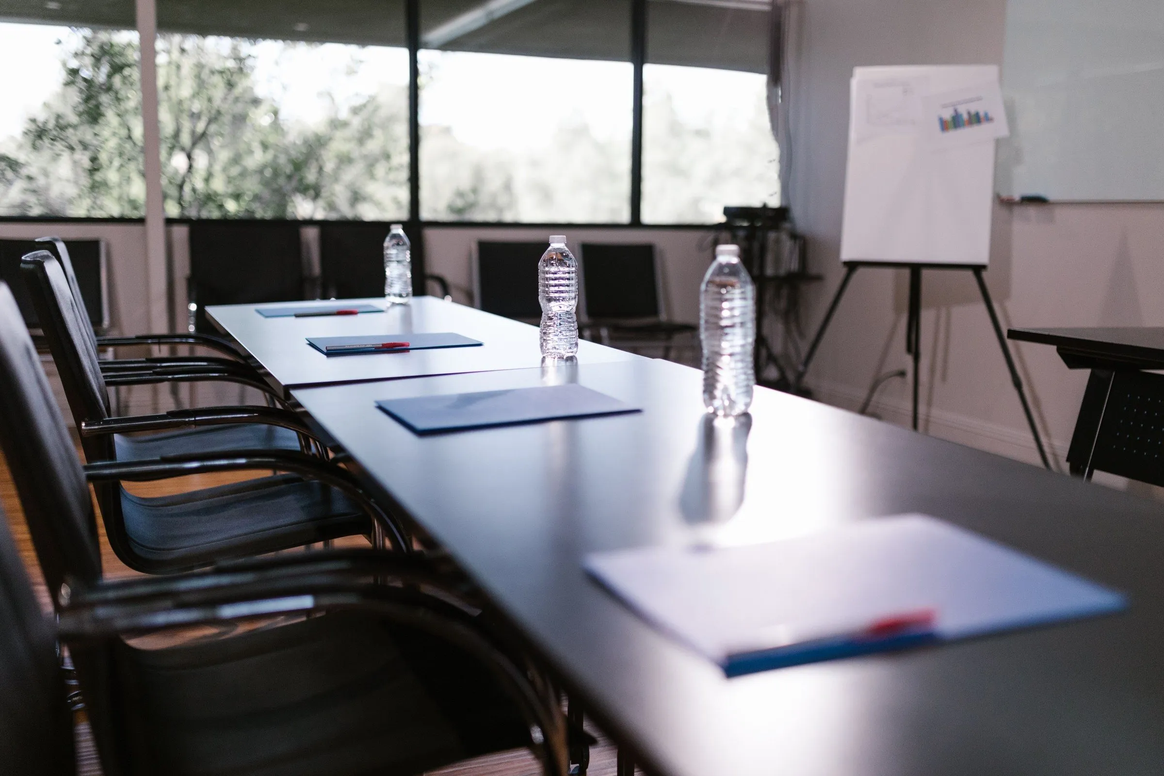 A meeting room set for a working session: blue folders and pens laid out along a long table, a flip chart by the window