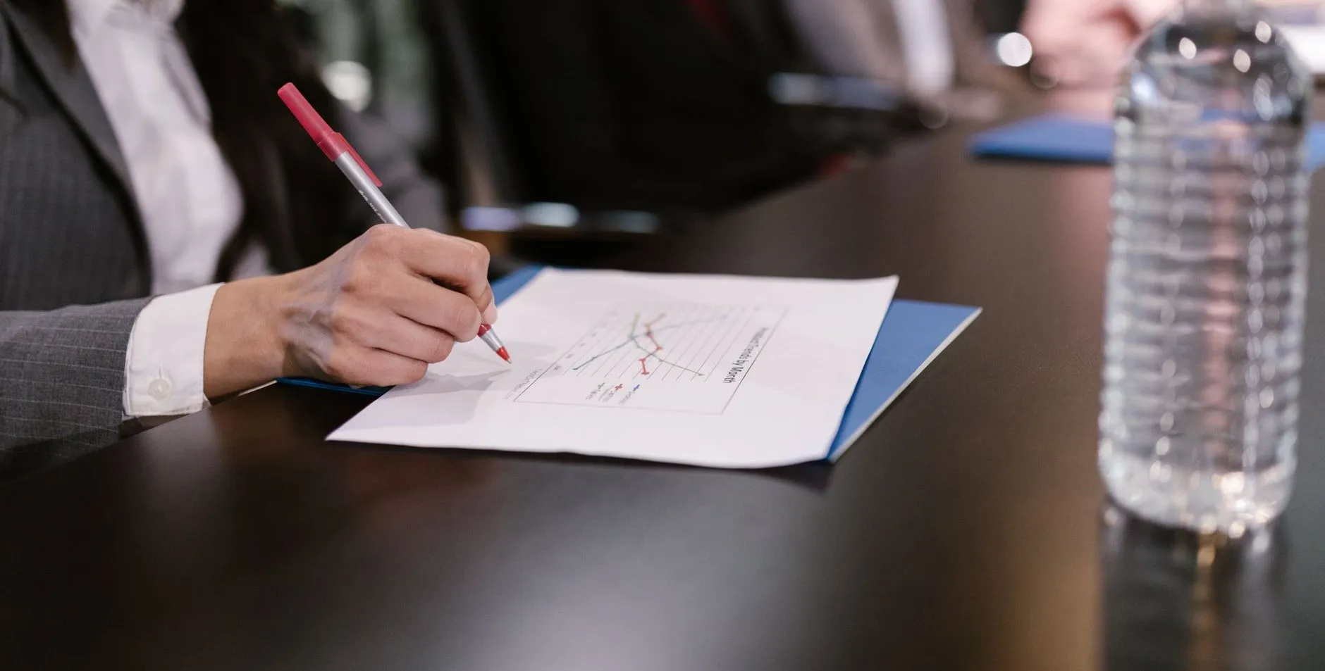 A hand marking a printed line chart at a long conference table, colleagues out of focus behind